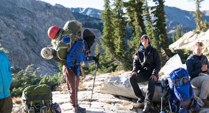 A person wearing a large backpack faces a person who is sitting on a rock and laughing. There are backpacks sitting on the ground, and two other people nearby. Behind them all, there are mountains. 